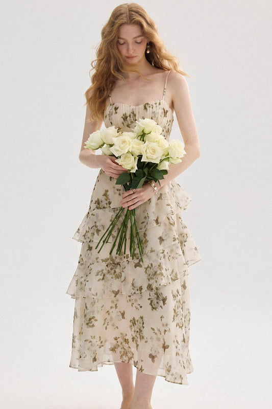 A woman wearing a sleeveless cream midi dress with a green floral print, holding a bouquet of white roses against a plain light gray background.