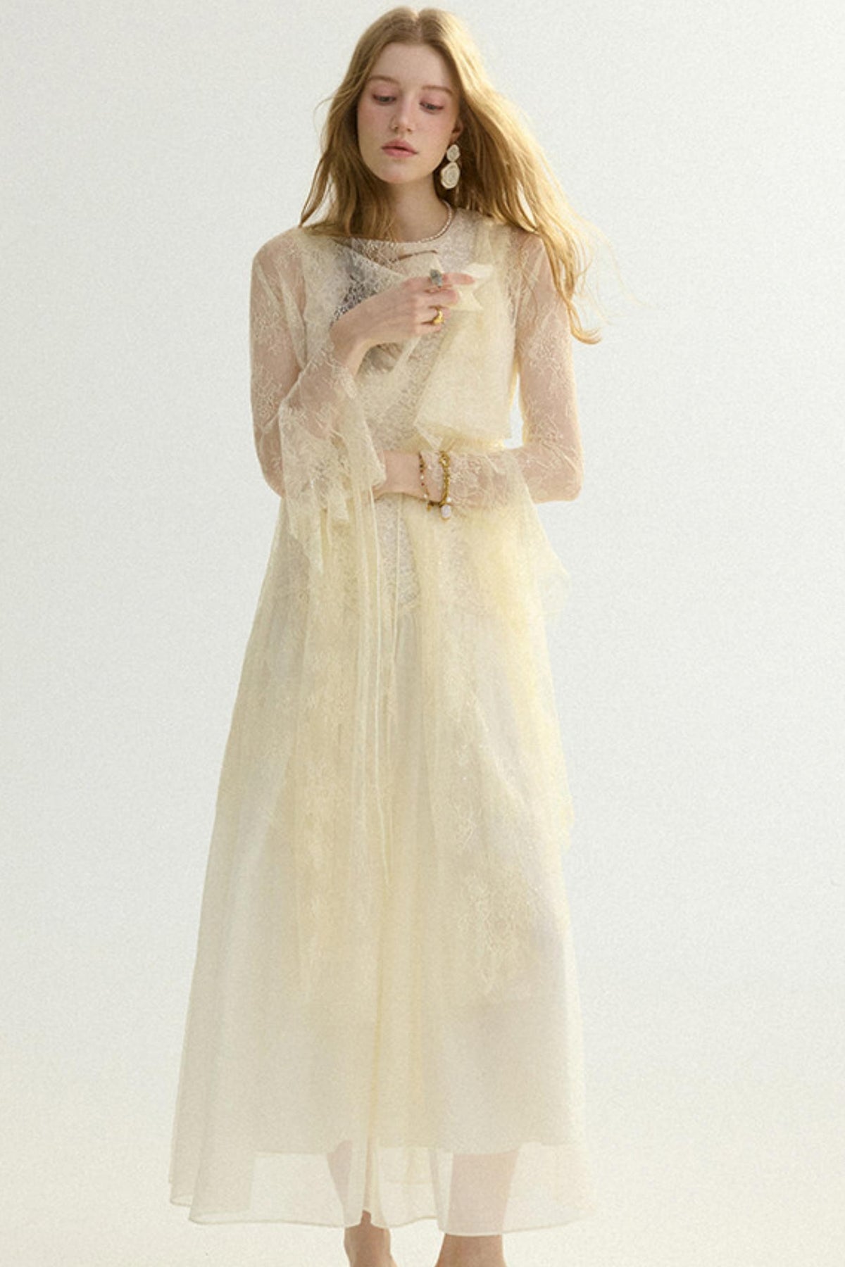 A woman wearing a long-sleeved cream-colored tulle dress with lace detailing, standing against a plain light background.