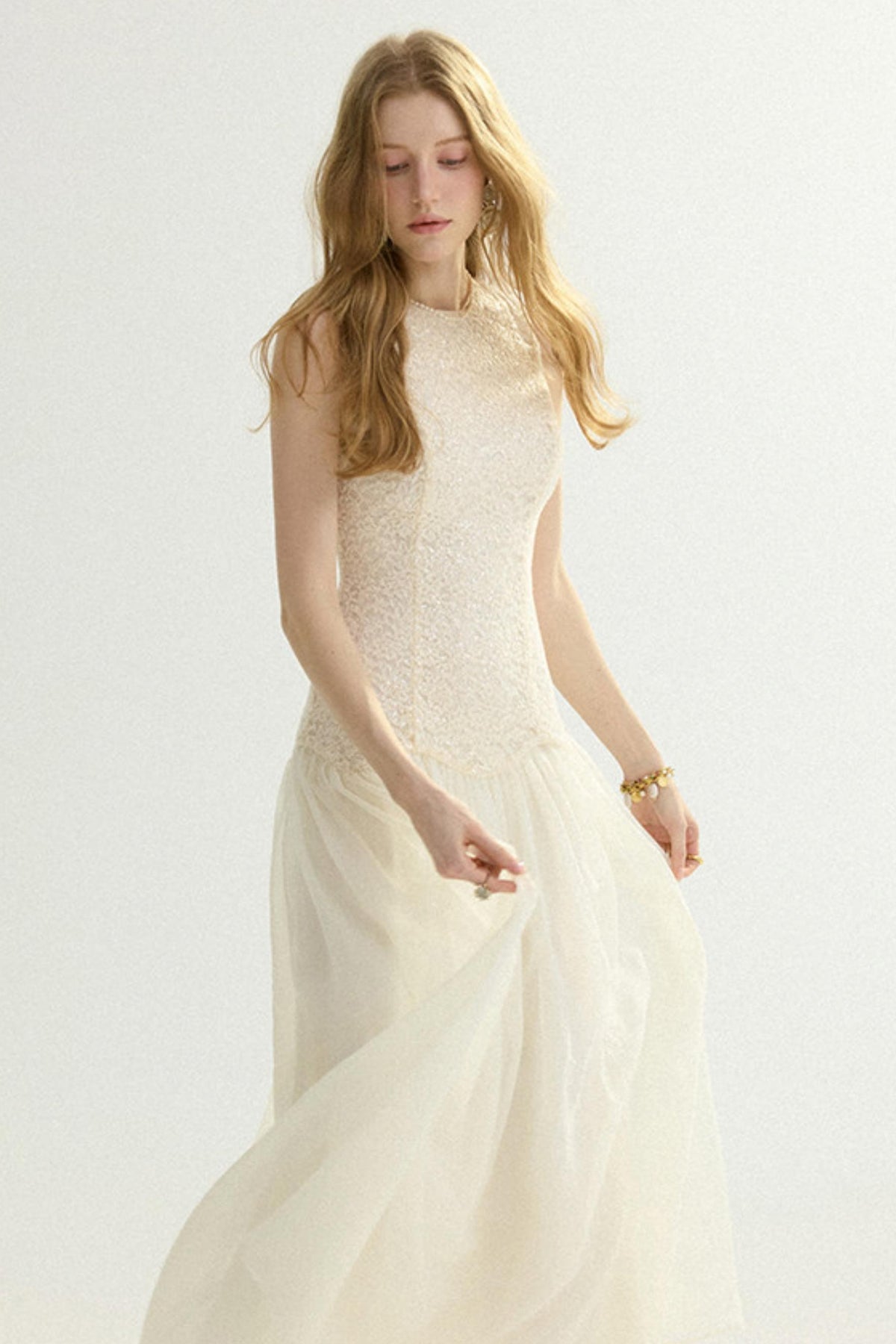 A woman wearing a sleeveless cream wedding dress with a textured lace bodice and a flowing tulle skirt, standing against a plain light background.
