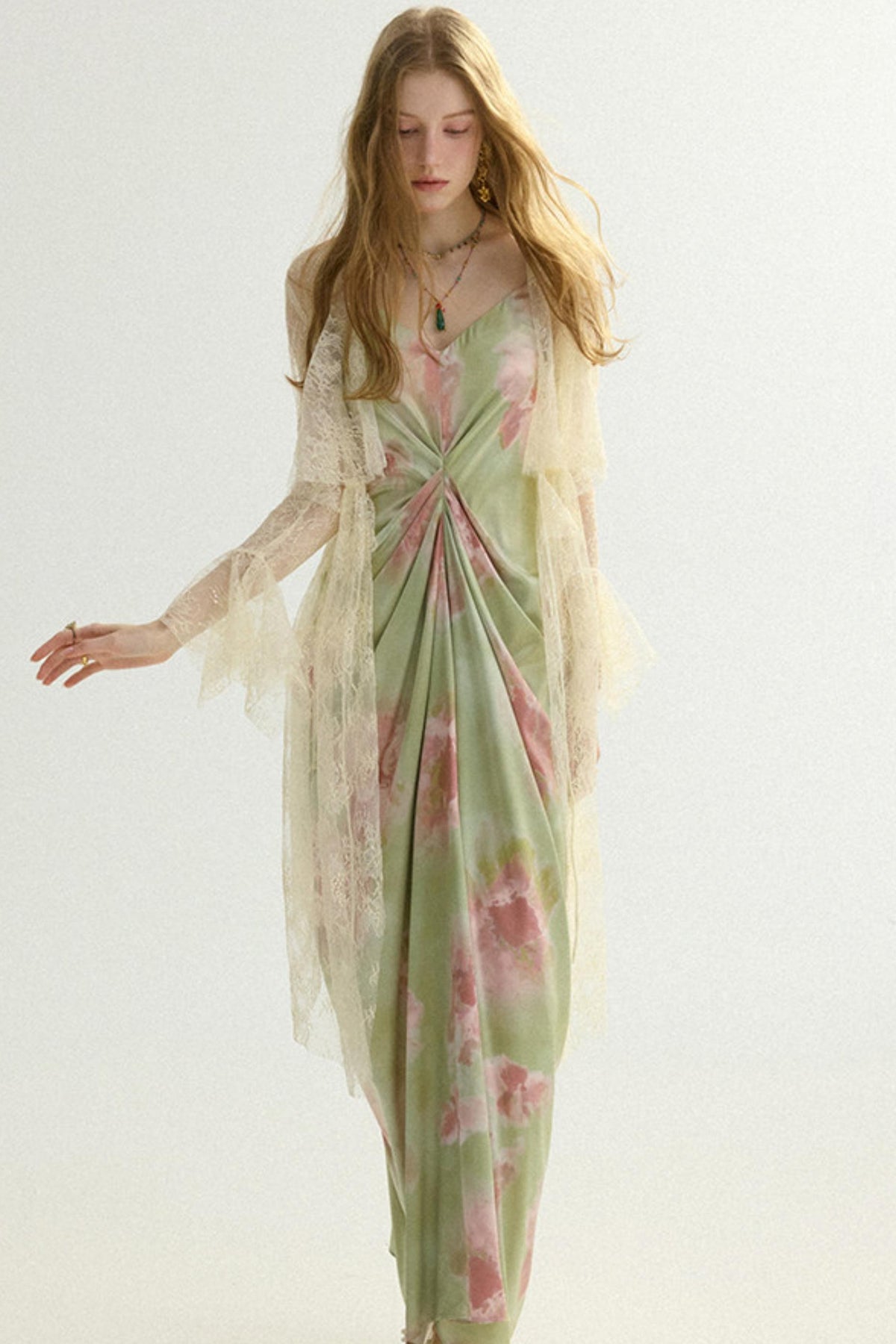 A woman modeling a long, flowing dress with a sheer overlay. The dress has a light sage green base with pink and white floral patterns, and the sheer overlay is a warm cream color.