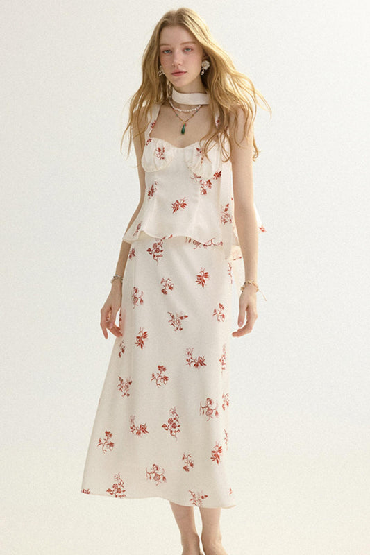 A woman modeling a sleeveless cream-colored dress with a red floral print, featuring a ruffled peplum waist and a matching midi skirt, set against a plain white studio background.