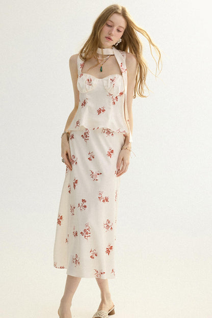 A woman modeling a sleeveless cream midi dress with a red floral print and a halter-style neckline with a small keyhole cutout, set against a plain white studio background.