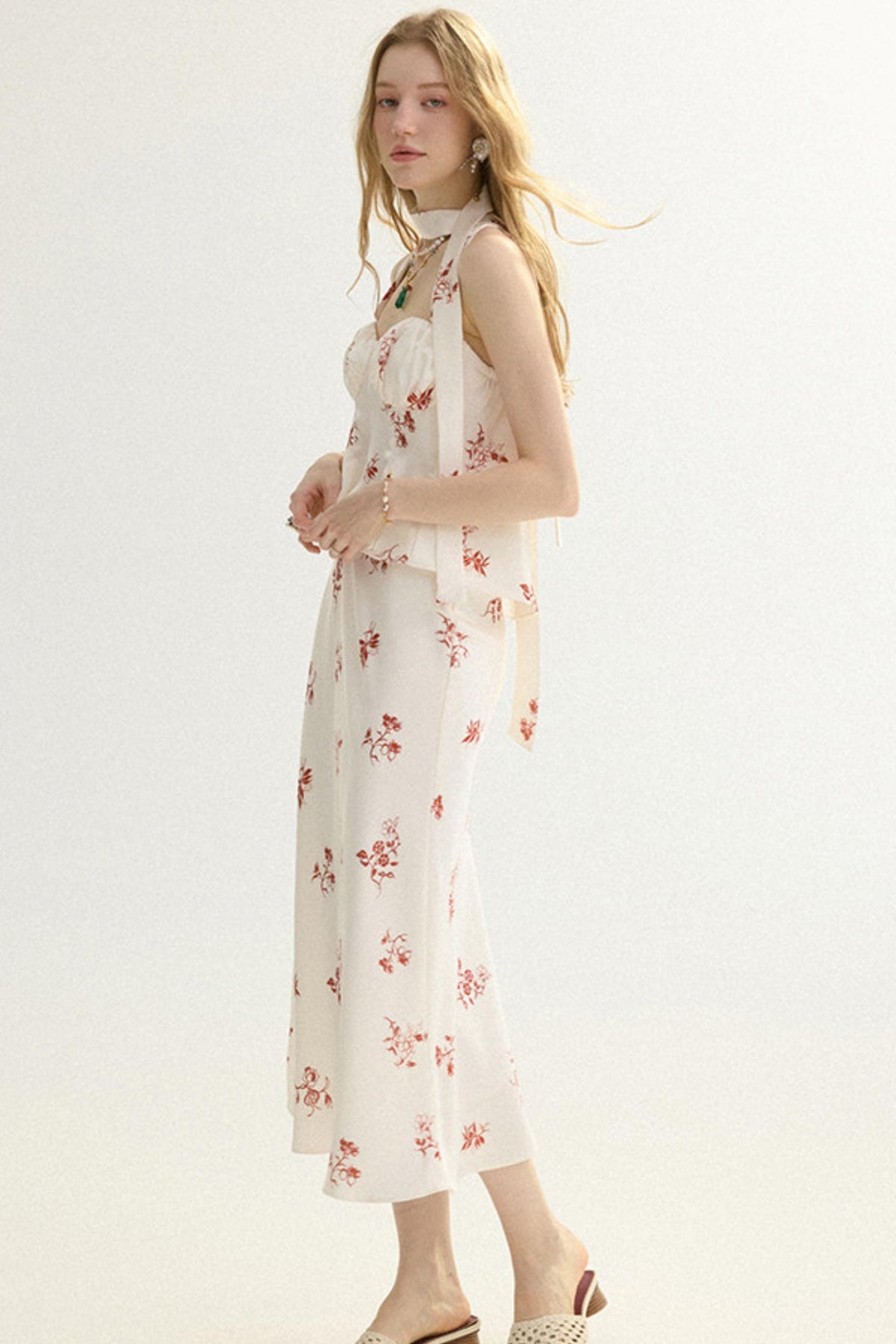 A woman modeling a sleeveless cream midi dress with a red and green floral print, featuring a high neck and tie details at the shoulders, set against a plain white studio background.
