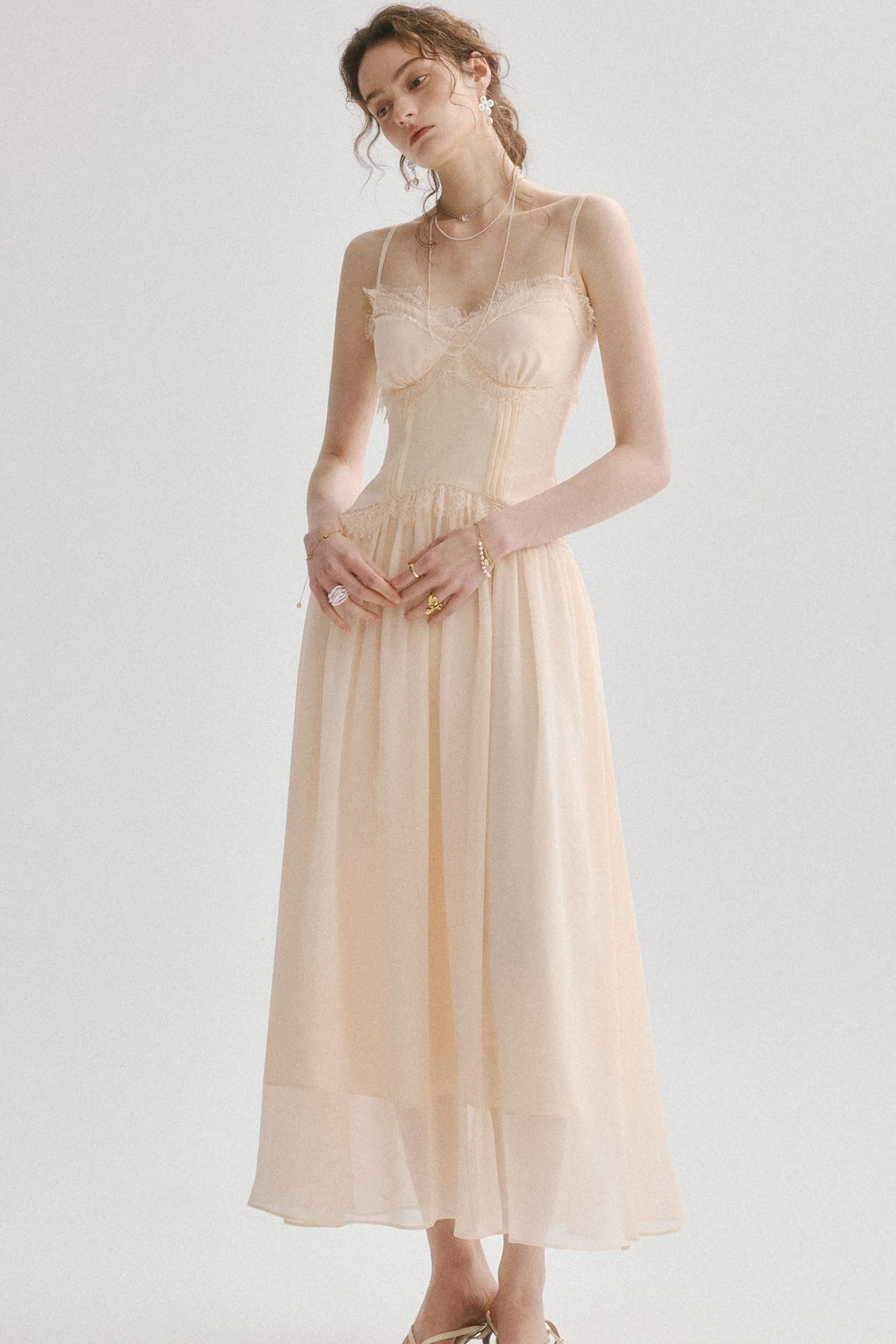 A woman modeling a light peach-beige midi dress with a structured lace corset bodice and a flowing chiffon skirt, set against a plain light gray studio background.