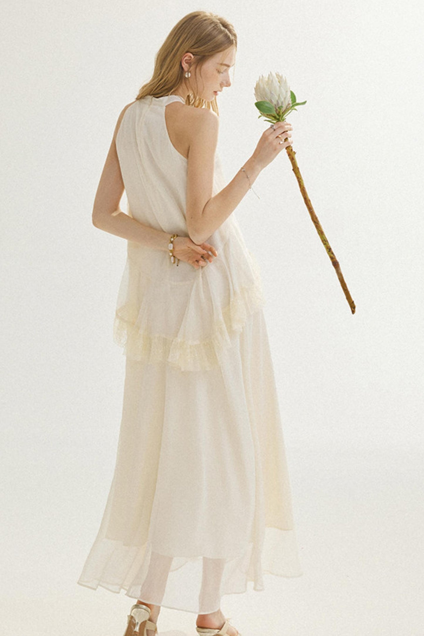 A woman stands against a plain light background wearing a long, flowing cream dress with a layered ruffle hem and holding a single white flower with a long stem.