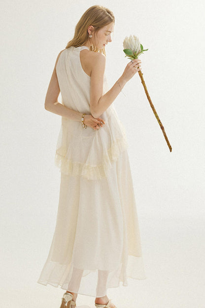 A woman stands against a plain light background wearing a long, flowing cream dress with a layered ruffle hem and holding a single white flower with a long stem.
