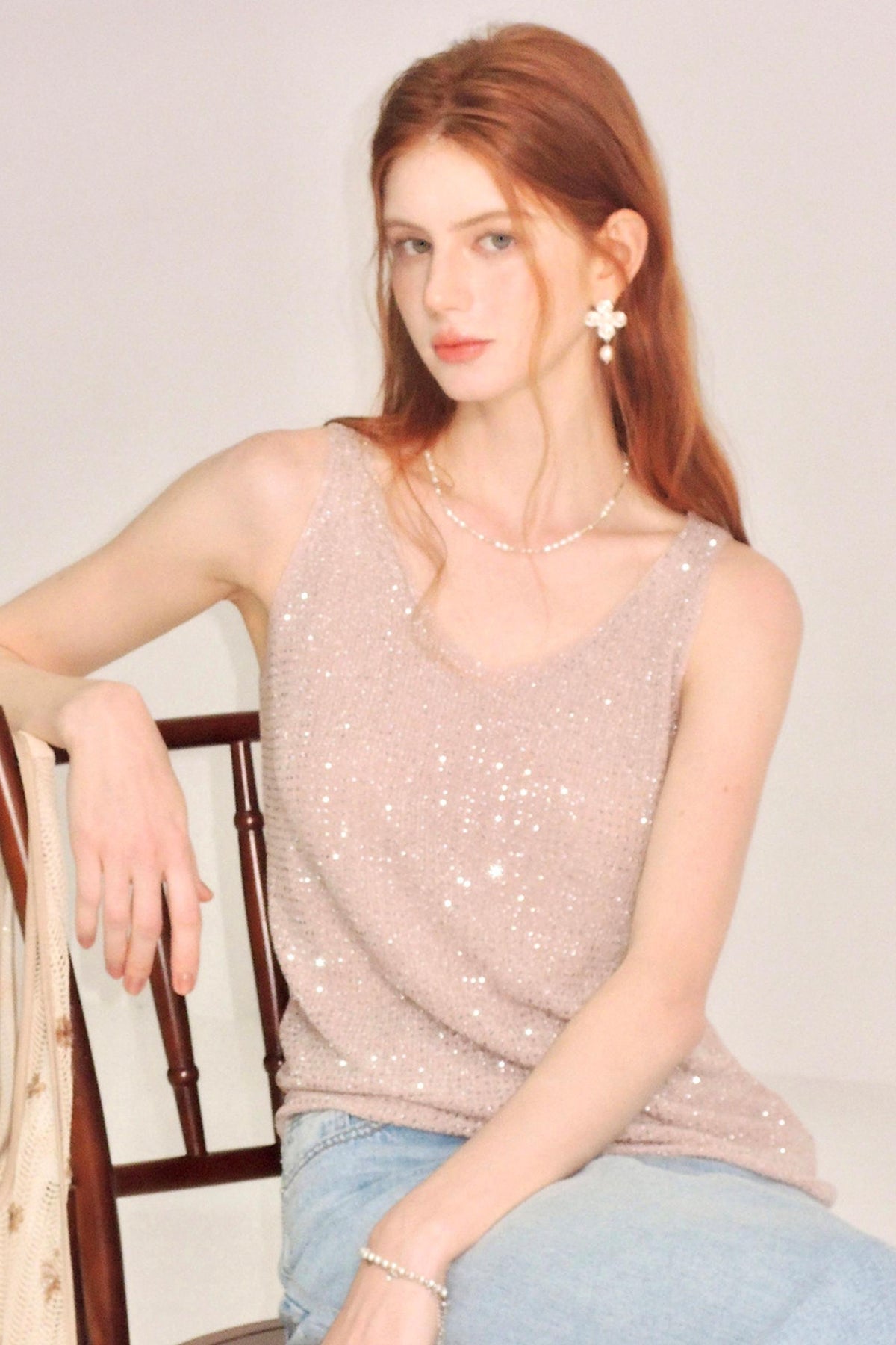 A woman with red hair wearing a sparkling light pink sequined sleeveless top and light blue jeans, sitting on a wooden chair against a plain light background.