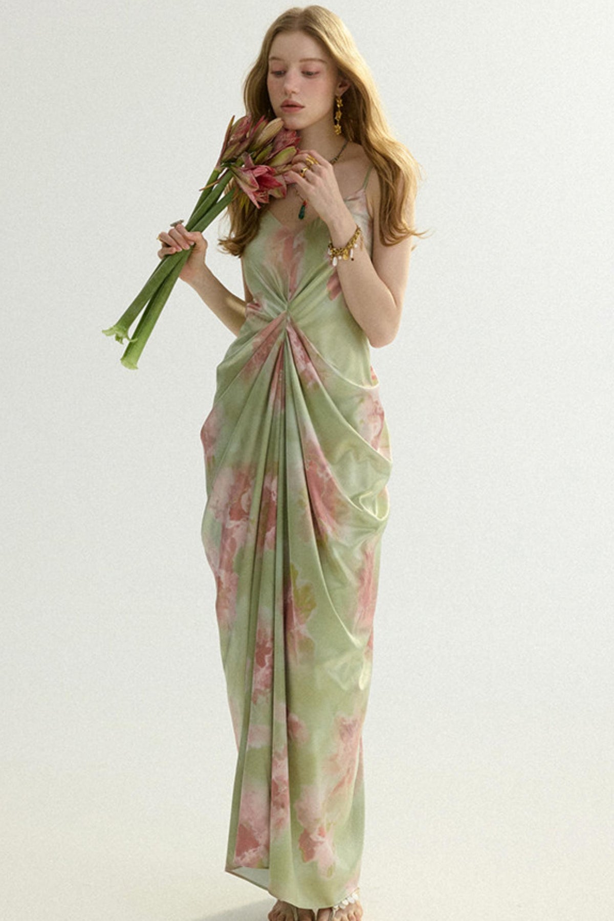 A woman modeling a long draped satin maxi dress in sage green with a pink floral print, holding a bouquet of pink flowers against a plain light background.