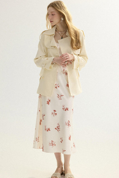 A woman modeling a cream-colored trench coat over a matching cream midi dress with a red floral print, paired with beige flat shoes against a plain white studio background.