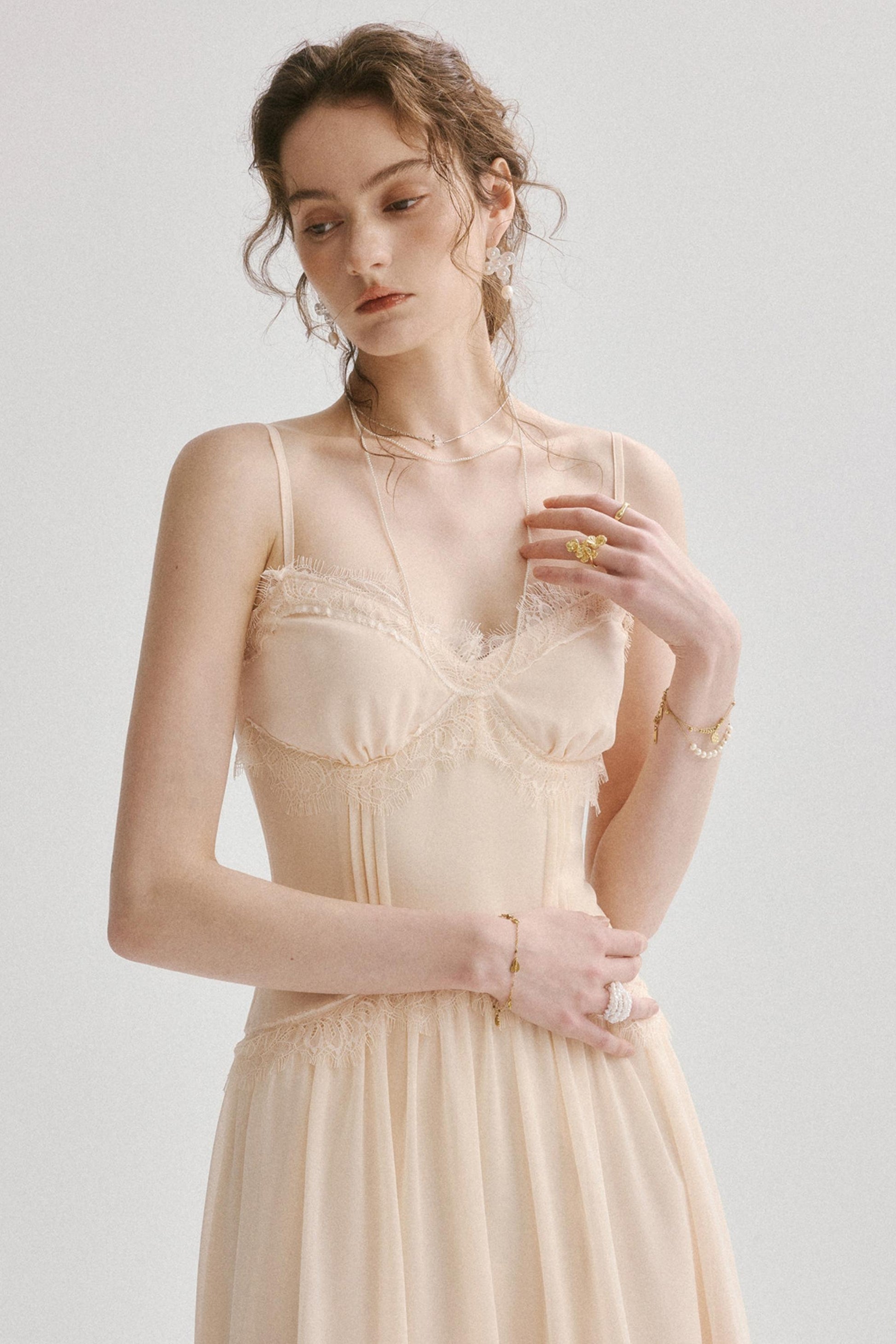 A woman wearing a light beige or cream-colored lace and mesh camisole dress with a pleated bodice and a flowing skirt, photographed against a plain light gray studio background.