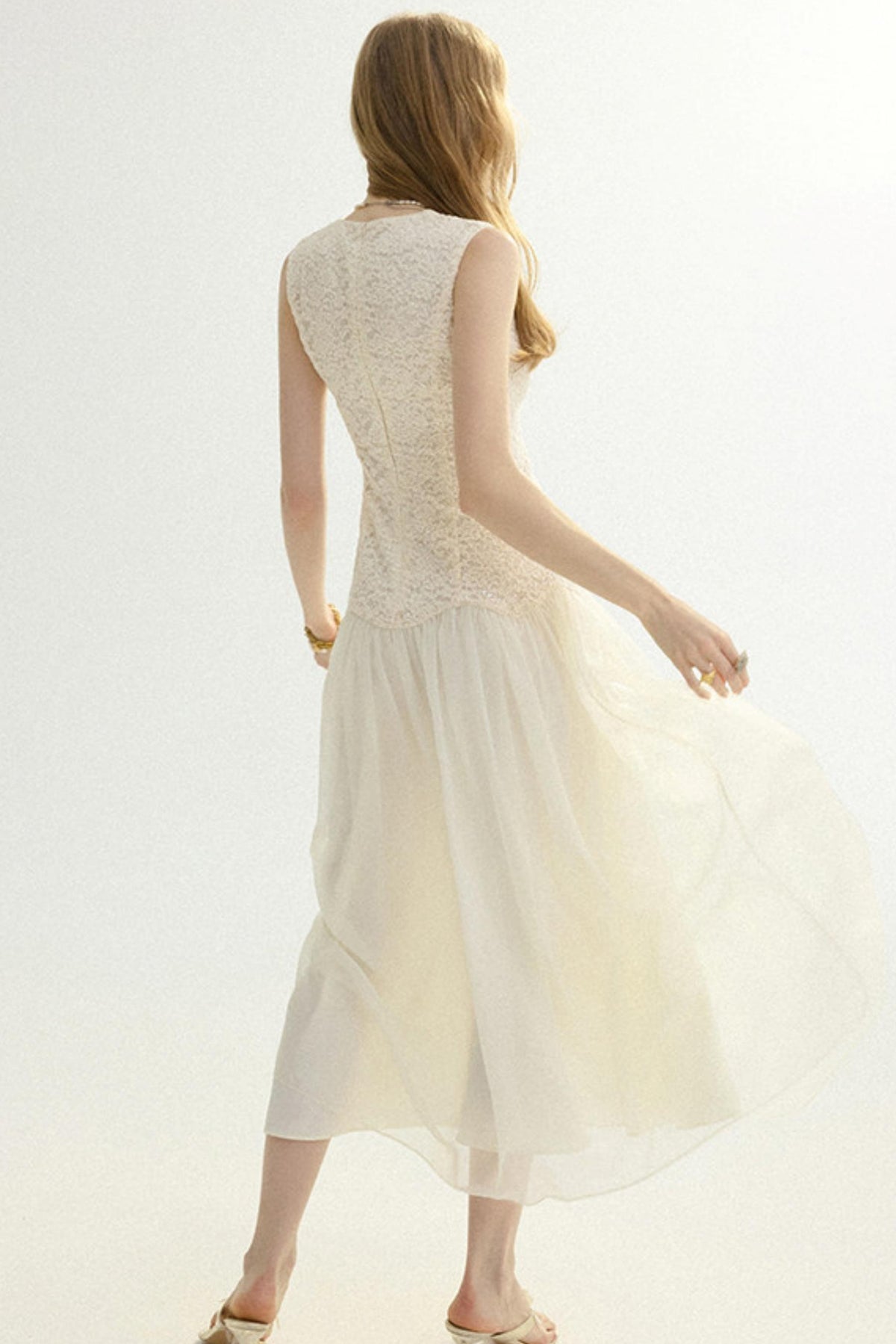A woman shown from the back wearing a sleeveless cream-colored midi dress with a lace bodice and flowing tulle skirt, set against a plain white studio background.