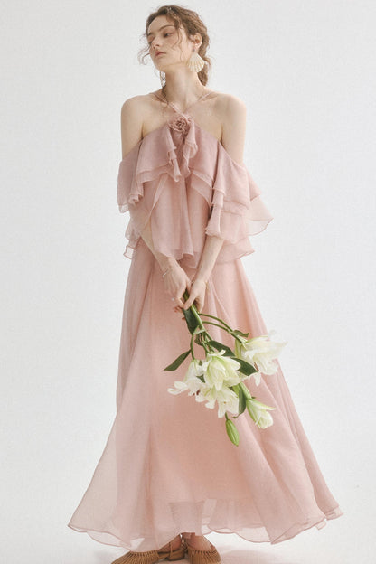 A woman wearing a dusty pink off-the-shoulder maxi dress with layered ruffle sleeves and a matching flowing skirt, holding a bouquet of white lilies against a plain light background.