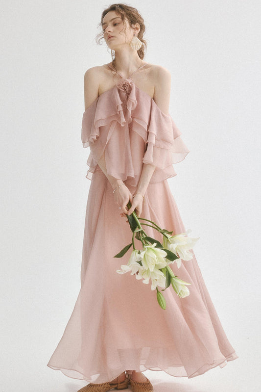 A woman wearing a dusty pink off-the-shoulder maxi dress with layered ruffle sleeves and a matching flowing skirt, holding a bouquet of white lilies against a plain light background.