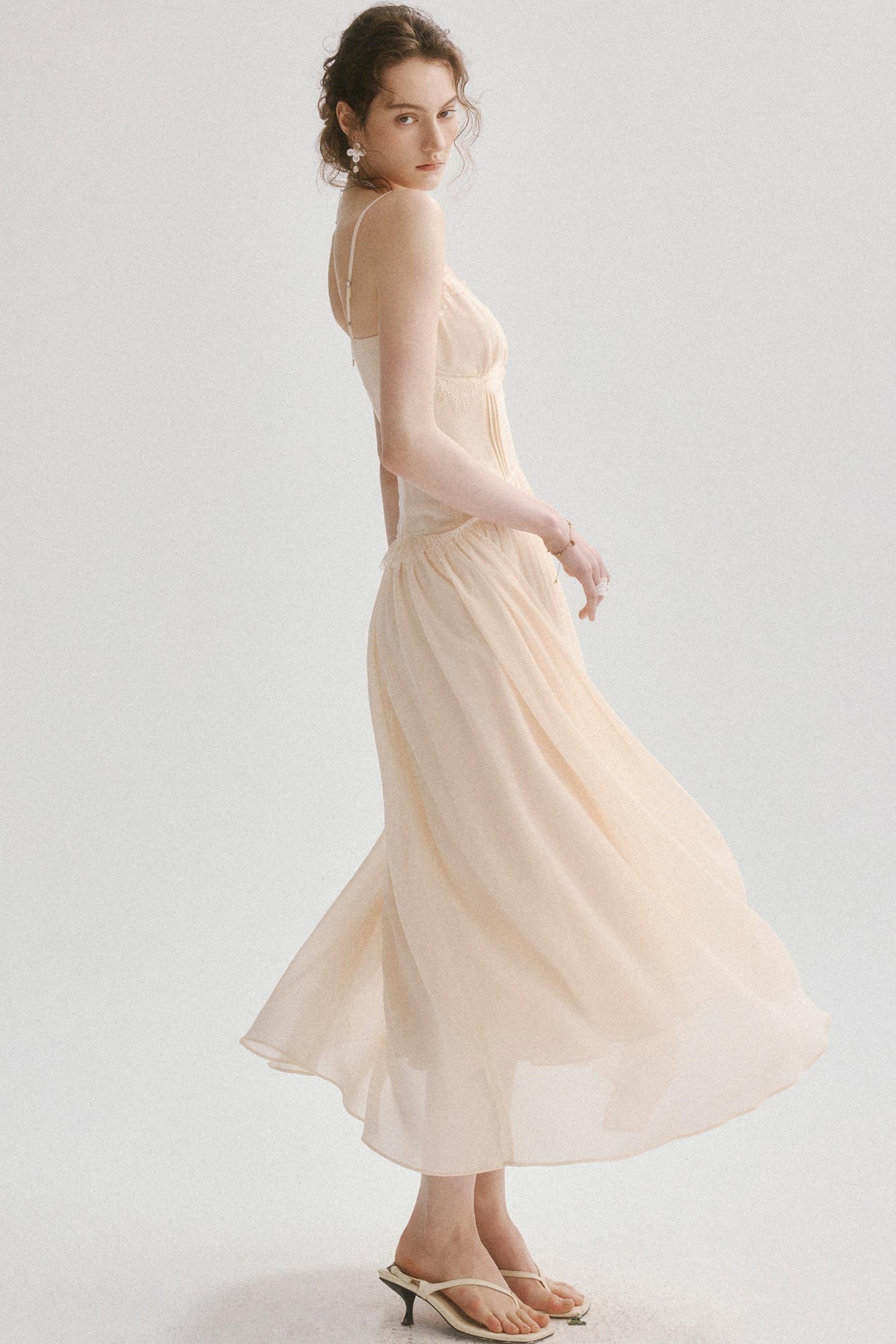 A woman wearing a flowing pale blush-beige maxi dress with a backless cutout, photographed against a clean light gray studio background.