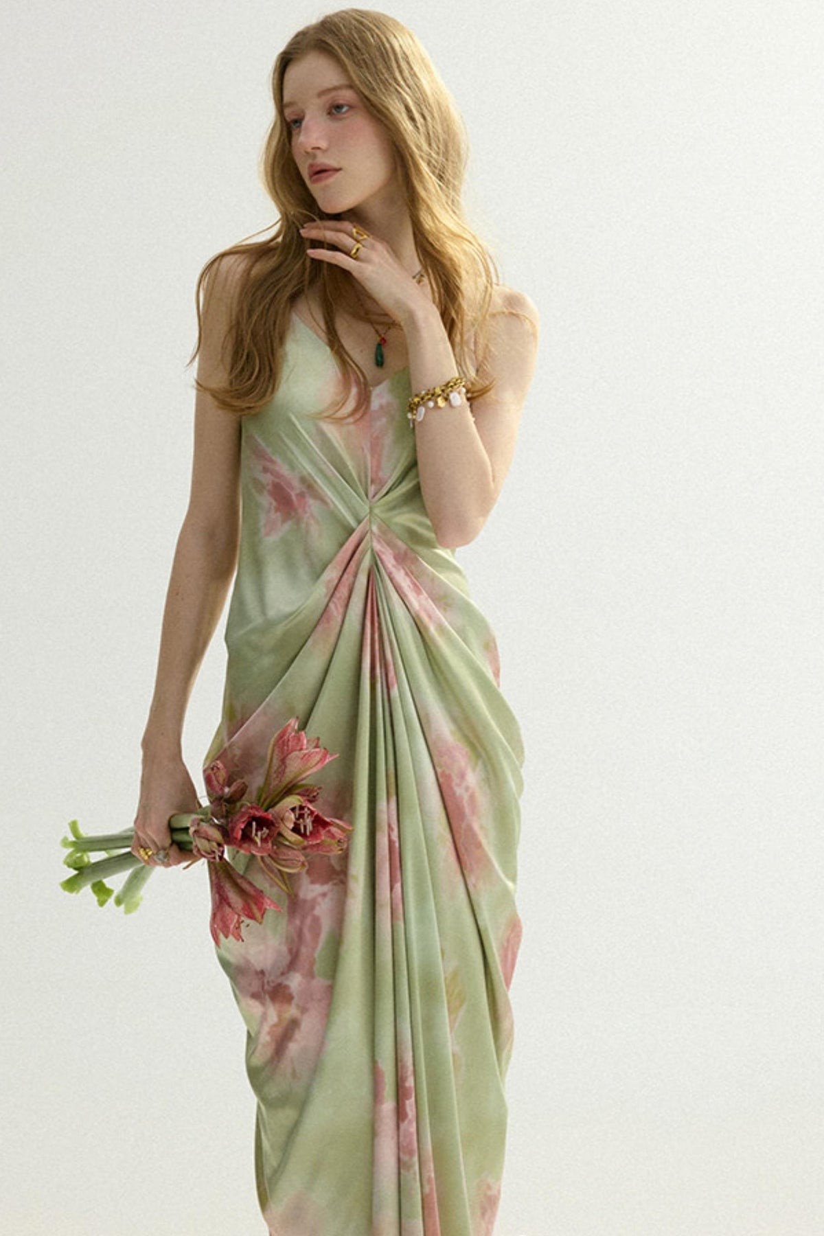 A woman modeling a light sage green sleeveless dress with a pink floral print and a twisted front detail, holding a small bouquet of pink flowers against a plain light background.