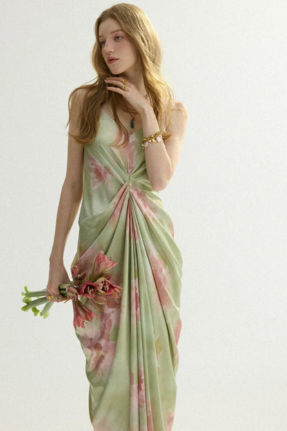 A woman modeling a light sage green sleeveless dress with a pink floral print and a twisted front detail, holding a small bouquet of pink flowers against a plain light background.