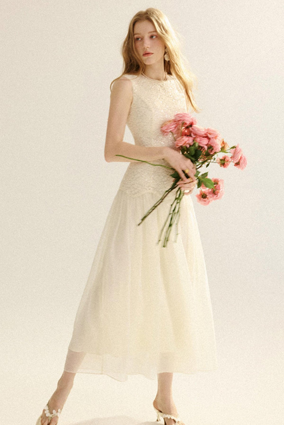 A woman modeling a sleeveless cream tulle midi dress with a lace bodice, holding a bouquet of pink roses against a plain light background.