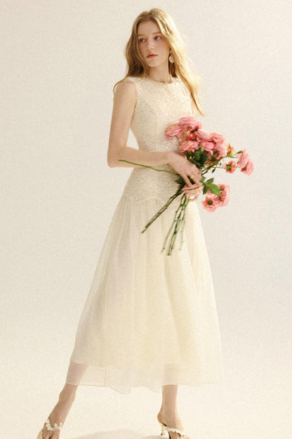 A woman modeling a sleeveless cream tulle midi dress with a lace bodice, holding a bouquet of pink roses against a plain light background.