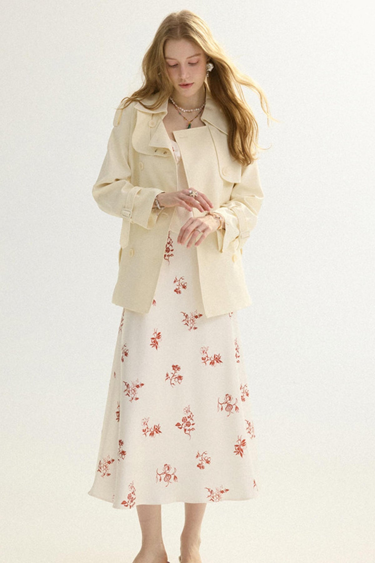 A woman modeling a cream-colored trench coat over a matching cream midi dress with a red floral pattern, set against a plain light background.