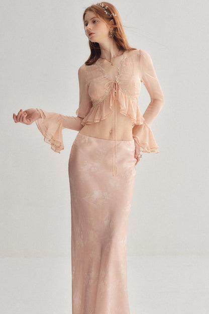 A woman modeling a two-piece peachy pink set consisting of a sheer long-sleeve crop top with ruffle details and a matching satin slip skirt, against a light gray studio background.