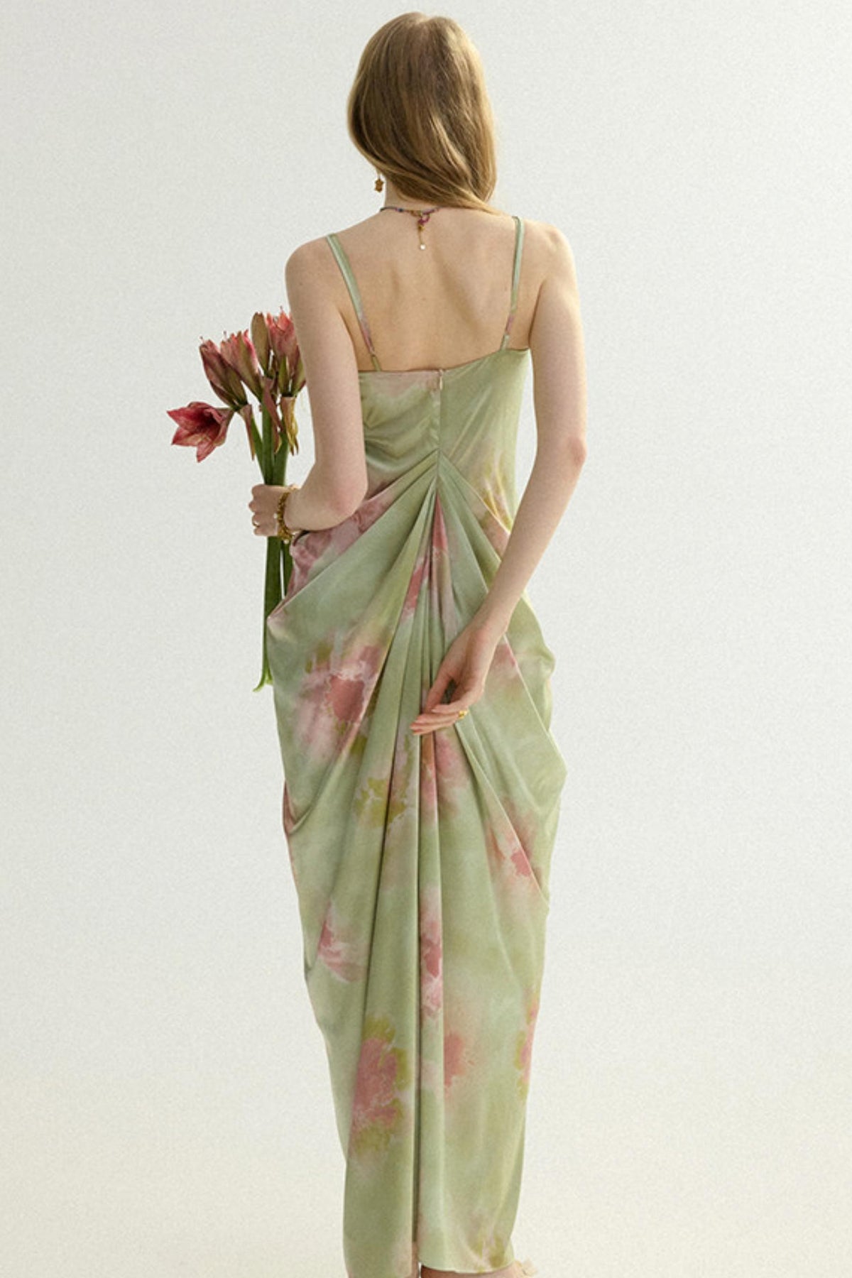 A woman stands with her back to the camera wearing a light sage green sleeveless maxi dress with a pink floral print and gathered draping at the waist. She is holding a small bouquet of pink flowers against a plain off-white studio background.