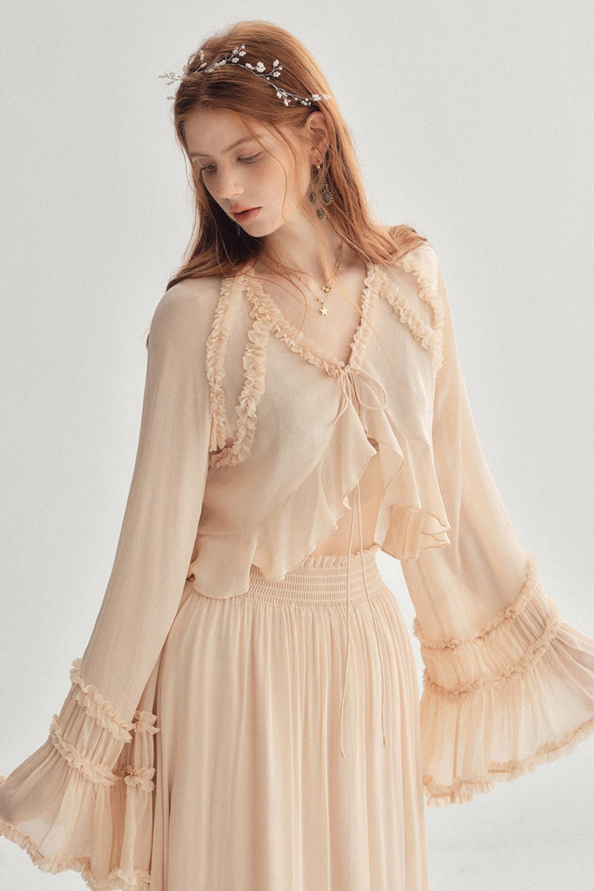 A woman wearing a light peach-beige bohemian-style two-piece set with a ruffled tie-neck blouse and matching pleated skirt, accessorized with a floral headband against a light gray studio background.