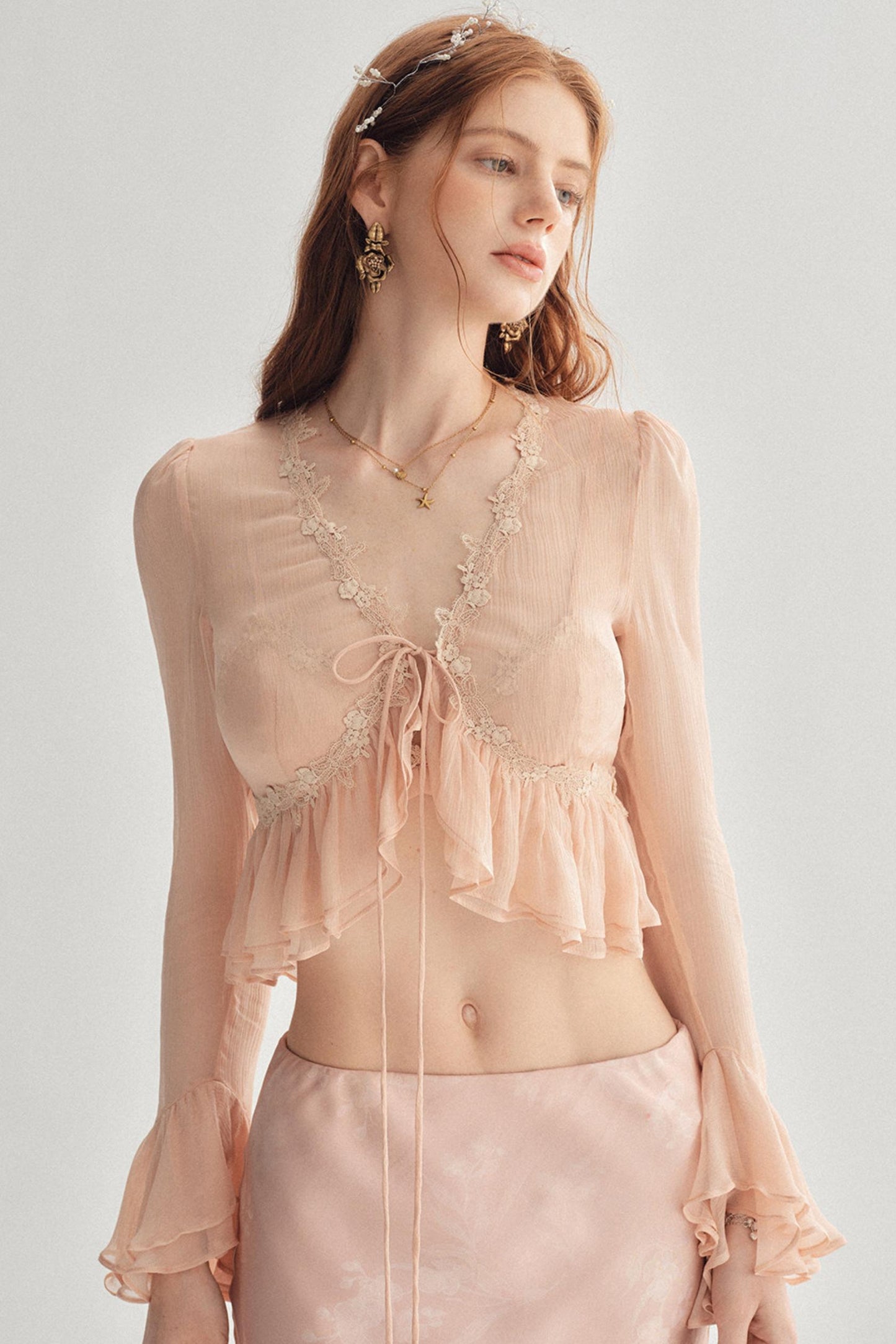 A woman modeling a light peach sheer long-sleeve crop top with lace trim, a front tie, and ruffled layers, paired with a matching satin skirt against a plain light gray studio background.