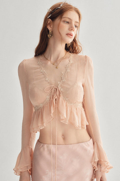 A woman modeling a light peach sheer long-sleeve crop top with lace trim, a front tie, and ruffled layers, paired with a matching satin skirt against a plain light gray studio background.