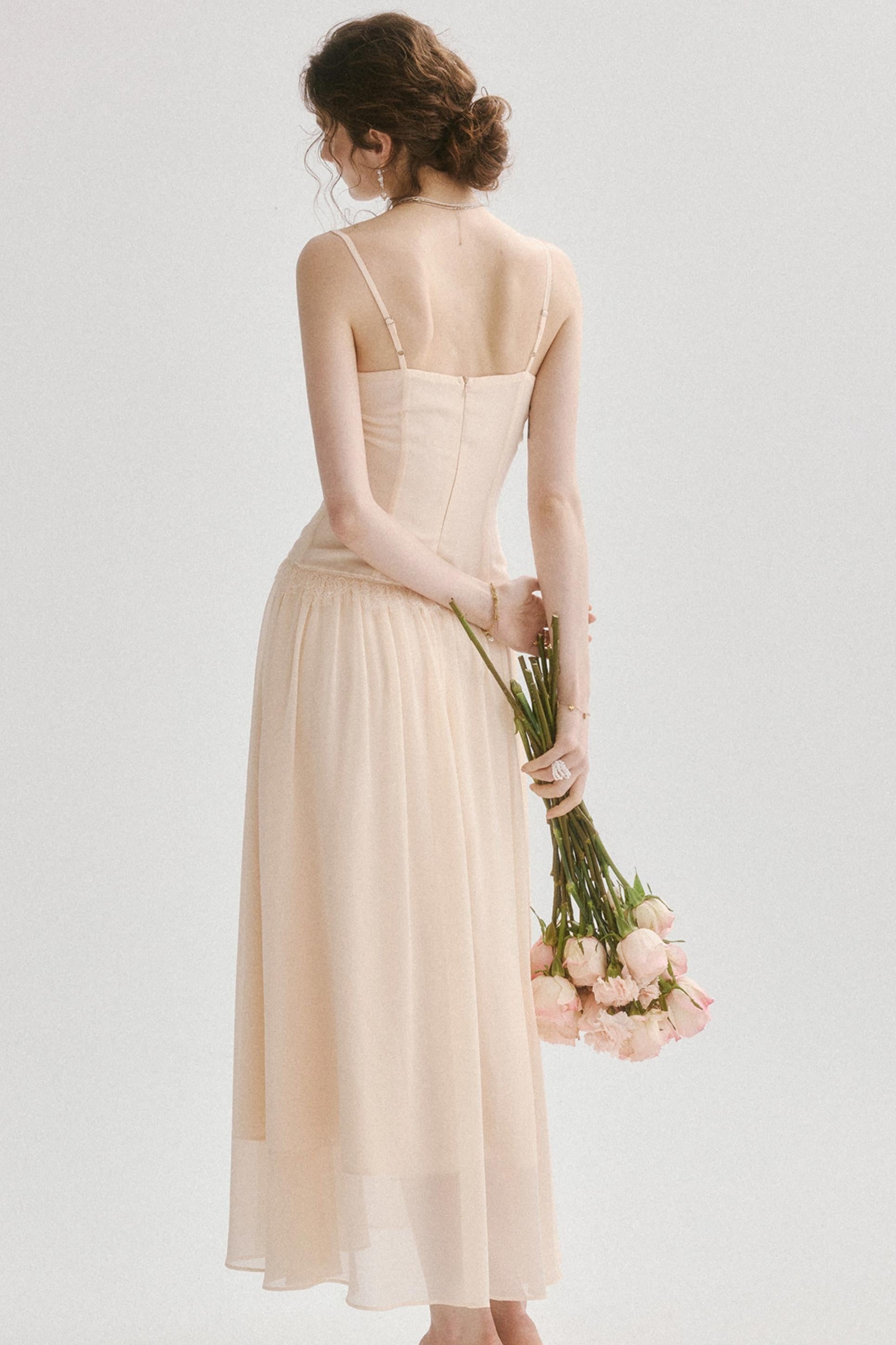 A woman shown from the back wearing a sleeveless, floor-length champagne dress and holding a bouquet of light pink roses against a plain light gray studio background.