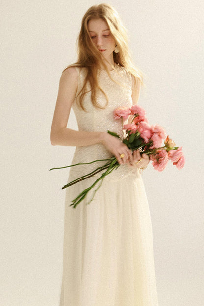 A woman wearing a sleeveless cream lace and tulle wedding dress, holding a bouquet of pink flowers against a plain light background.