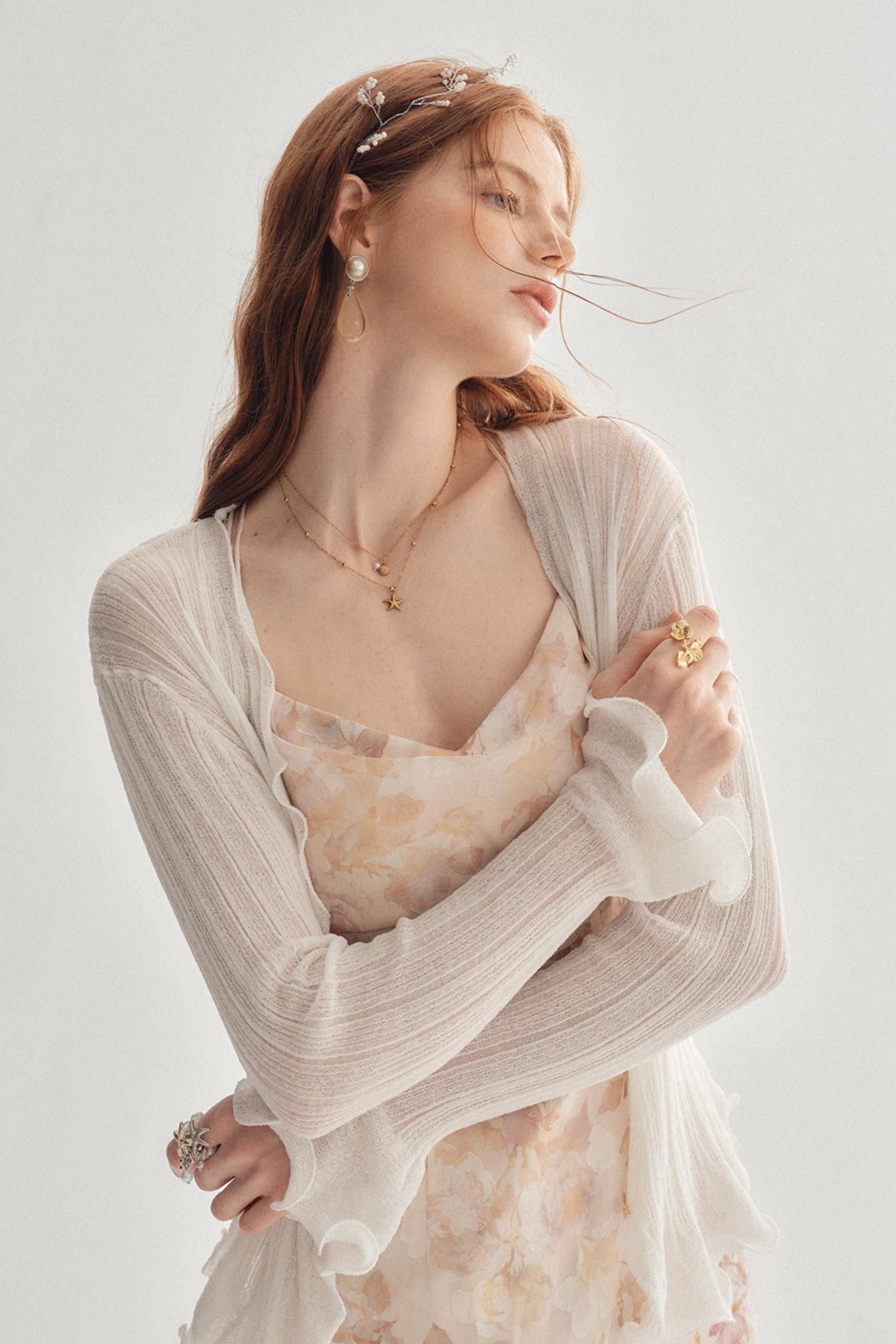 A woman wearing a light beige open-front cardigan over a pale floral dress, styled with delicate gold jewelry and a silver headpiece against a soft light gray background.