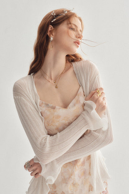 A woman wearing a light beige open-front cardigan over a pale floral dress, styled with delicate gold jewelry and a silver headpiece against a soft light gray background.