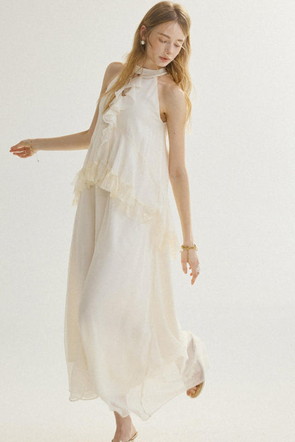 A woman modeling a sleeveless, high-neck, flowing maxi dress in a warm off-white or cream color, set against a plain light background.