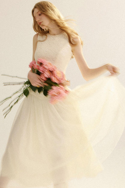 A woman wearing a sleeveless cream tulle wedding dress with a textured bodice, holding a bouquet of pink roses against a plain light background.
