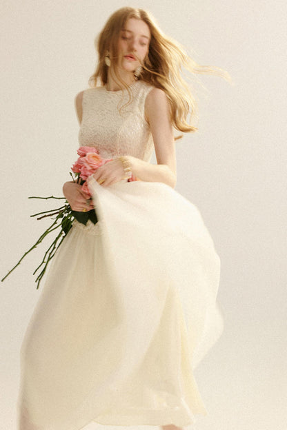A woman wearing a sleeveless cream lace bodice and flowing tulle skirt dress, holding a bouquet of pink roses against a plain light background.