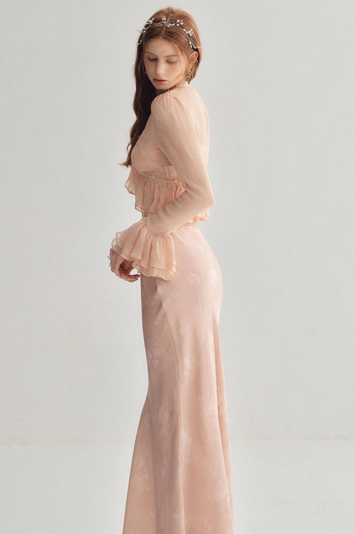 A woman wearing a long, sheer blush pink gown with ruffled details and a matching headpiece, photographed against a plain light gray studio background.