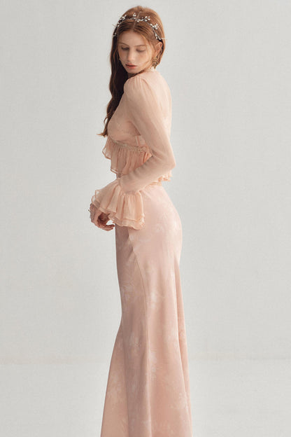 A woman wearing a long, sheer blush pink gown with ruffled details and a matching headpiece, photographed against a plain light gray studio background.