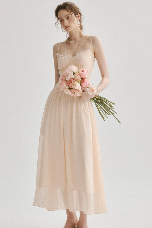 A woman wearing a sleeveless, flowing champagne-colored midi dress and holding a bouquet of pink flowers, photographed against a plain light gray studio background.