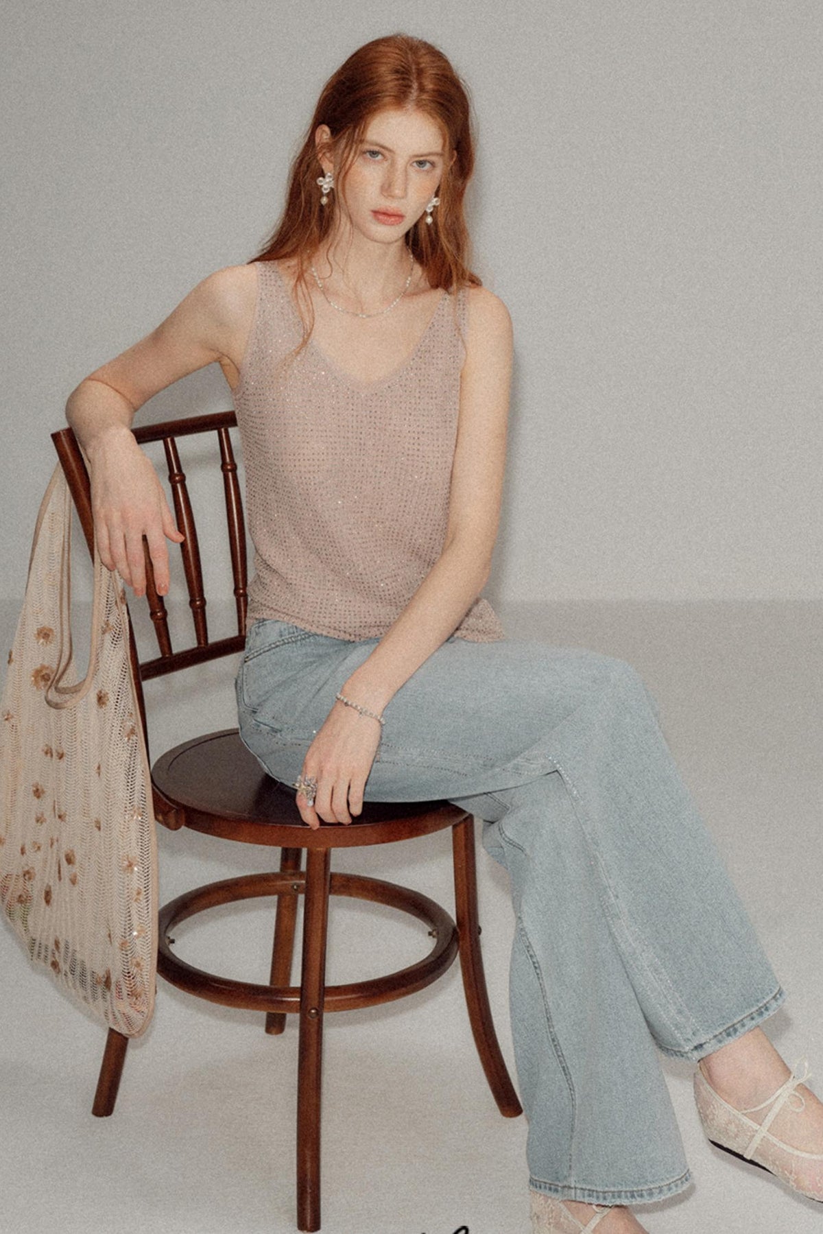 A woman with red hair sits on a wooden chair wearing a beige knit sleeveless top and light blue wide-leg jeans, holding a sheer beige patterned bag against a plain light gray studio background.