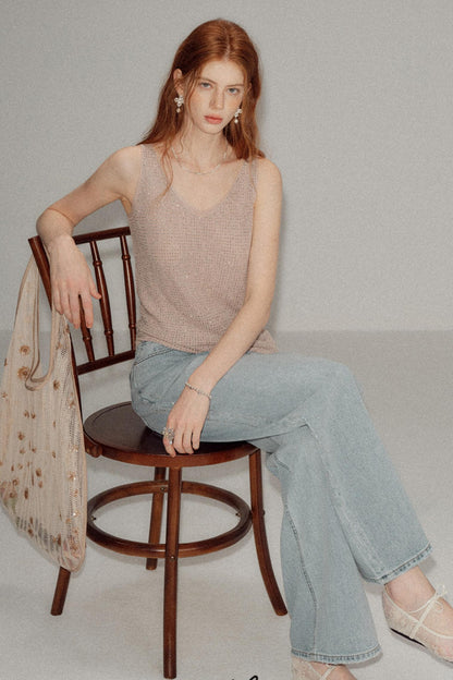 A woman with red hair sits on a wooden chair wearing a beige knit sleeveless top and light blue wide-leg jeans, holding a sheer beige patterned bag against a plain light gray studio background.