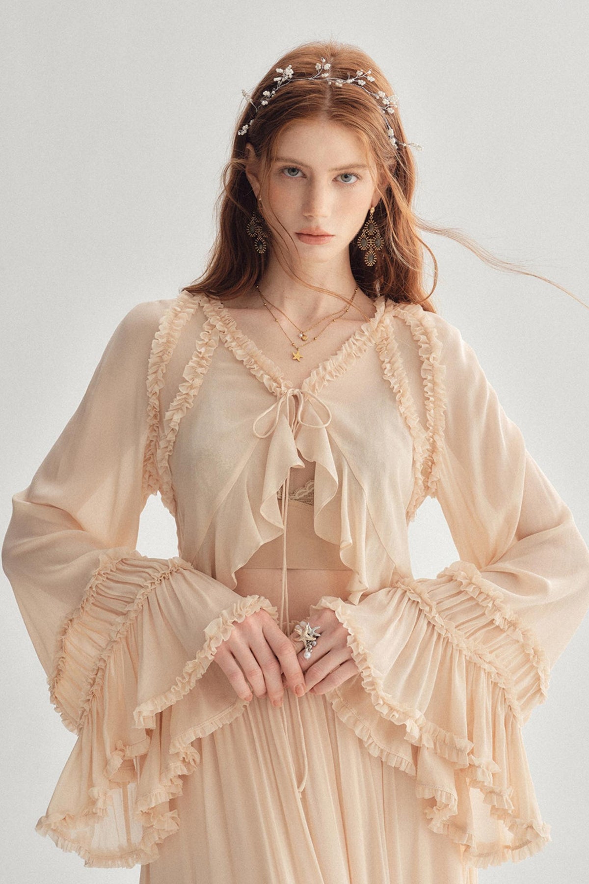 A woman wearing a light beige bohemian-style two-piece outfit with ruffled trim, long bell sleeves, and a matching headpiece, photographed against a plain light gray studio background.