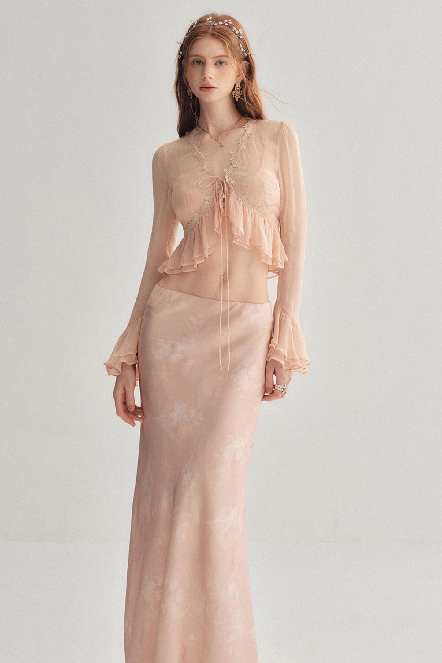 A woman modeling a two-piece sheer blush pink outfit against a light gray studio background. The set includes a long-sleeve ruffled crop top with a front tie and a matching long skirt.