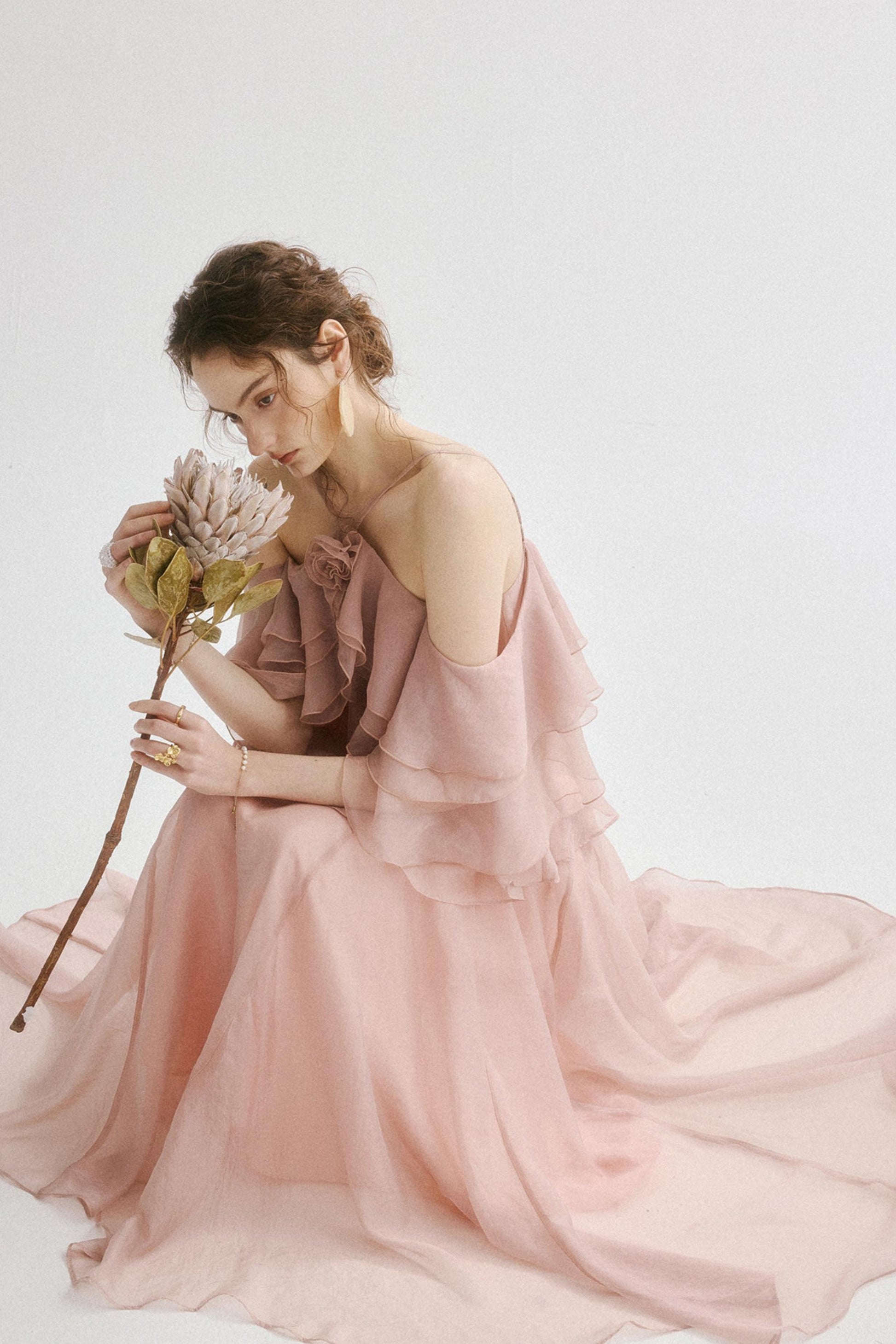 A woman in a flowing pale pink off-the-shoulder gown sits on a white studio floor, holding a dried protea flower.