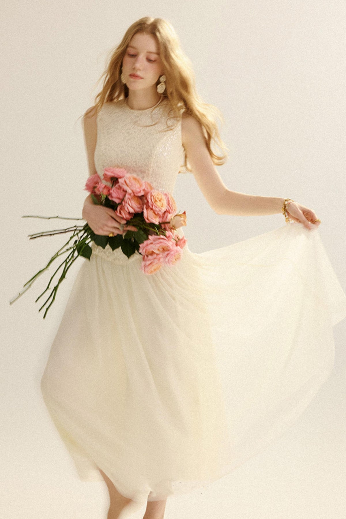 A woman wearing a sleeveless cream tulle dress with a textured lace bodice, holding a bouquet of pink roses and lifting the skirt to show its flow, against a plain light background.