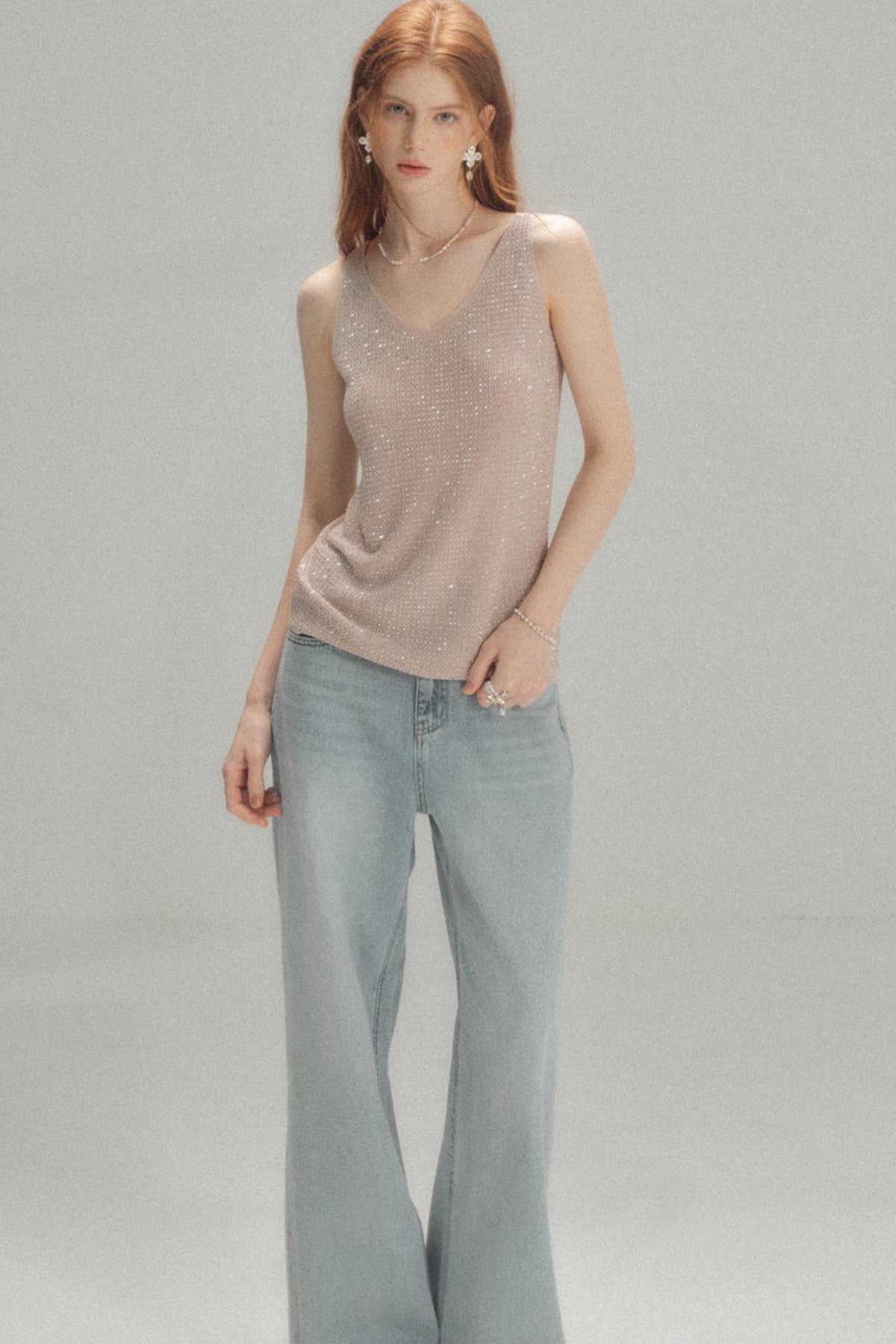 A model with red hair wearing a shimmering light beige sleeveless knit top and light blue wide-leg jeans, standing against a neutral gray studio background.