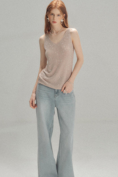 A model with red hair wearing a shimmering light beige sleeveless knit top and light blue wide-leg jeans, standing against a neutral gray studio background.