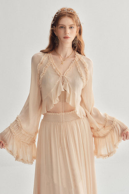 A woman modeling a light beige bohemian-style two-piece set consisting of a sheer long-sleeve crop top with lace trim and tie-front detail, paired with a matching pleated maxi skirt, against a plain light gray background.