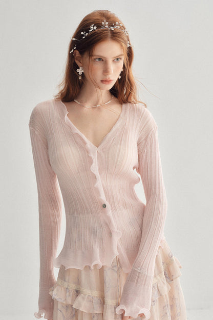 A woman wearing a light pink long-sleeve ribbed cardigan with ruffled trim over a matching pale floral dress, styled with a floral headband and delicate jewelry against a plain light gray background.
