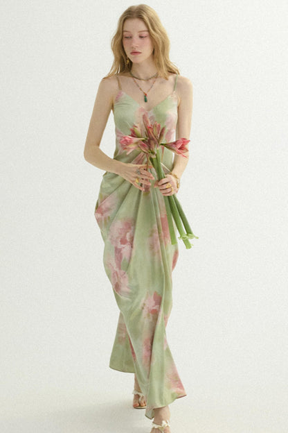 A woman modeling a light sage green spaghetti-strap maxi dress with a pink floral print, holding a bouquet of pink flowers against a plain white studio background.
