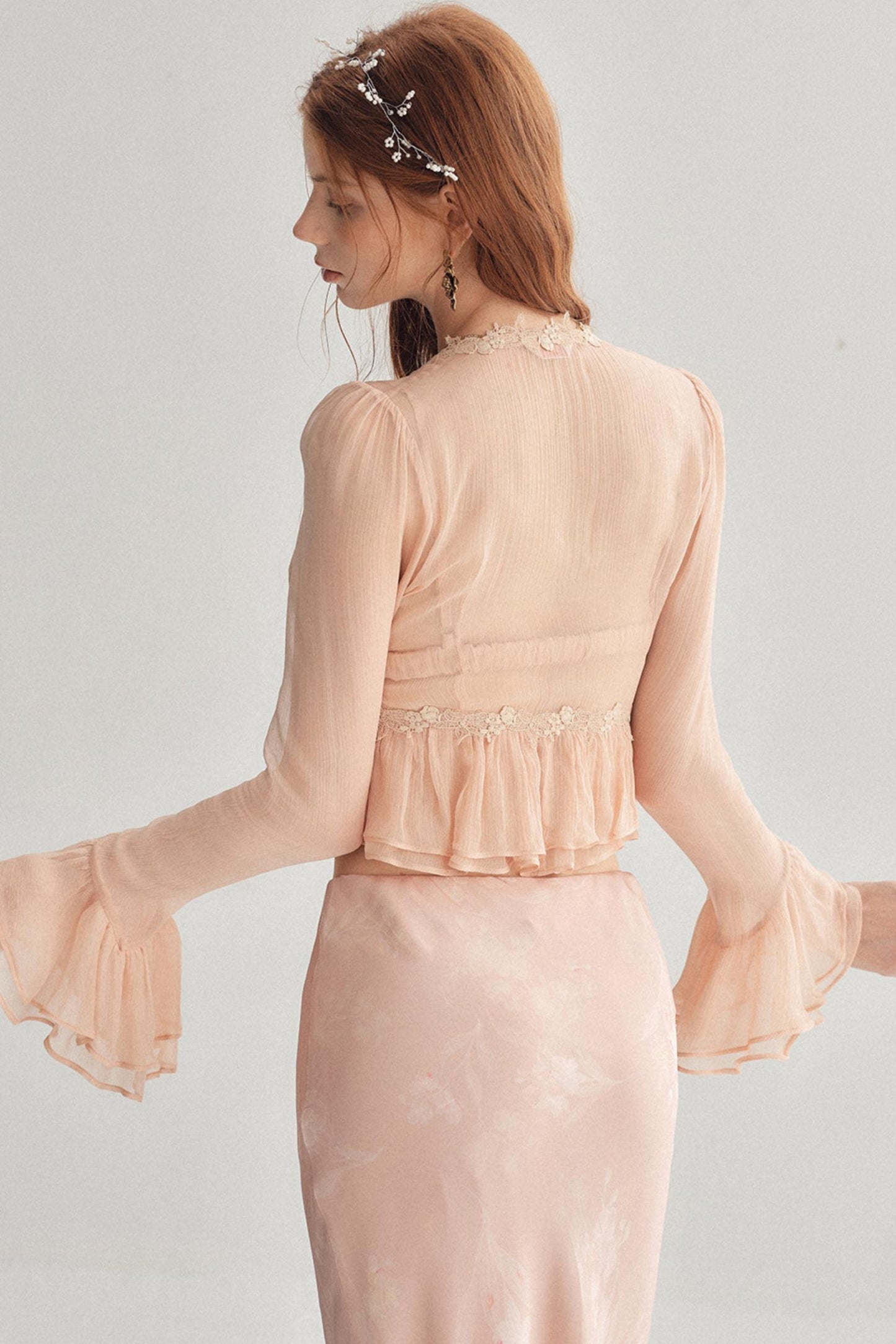 A woman shown from the back wearing a pale blush pink long-sleeve dress with a ruffled waistband and flared bell sleeves, against a light gray studio background.